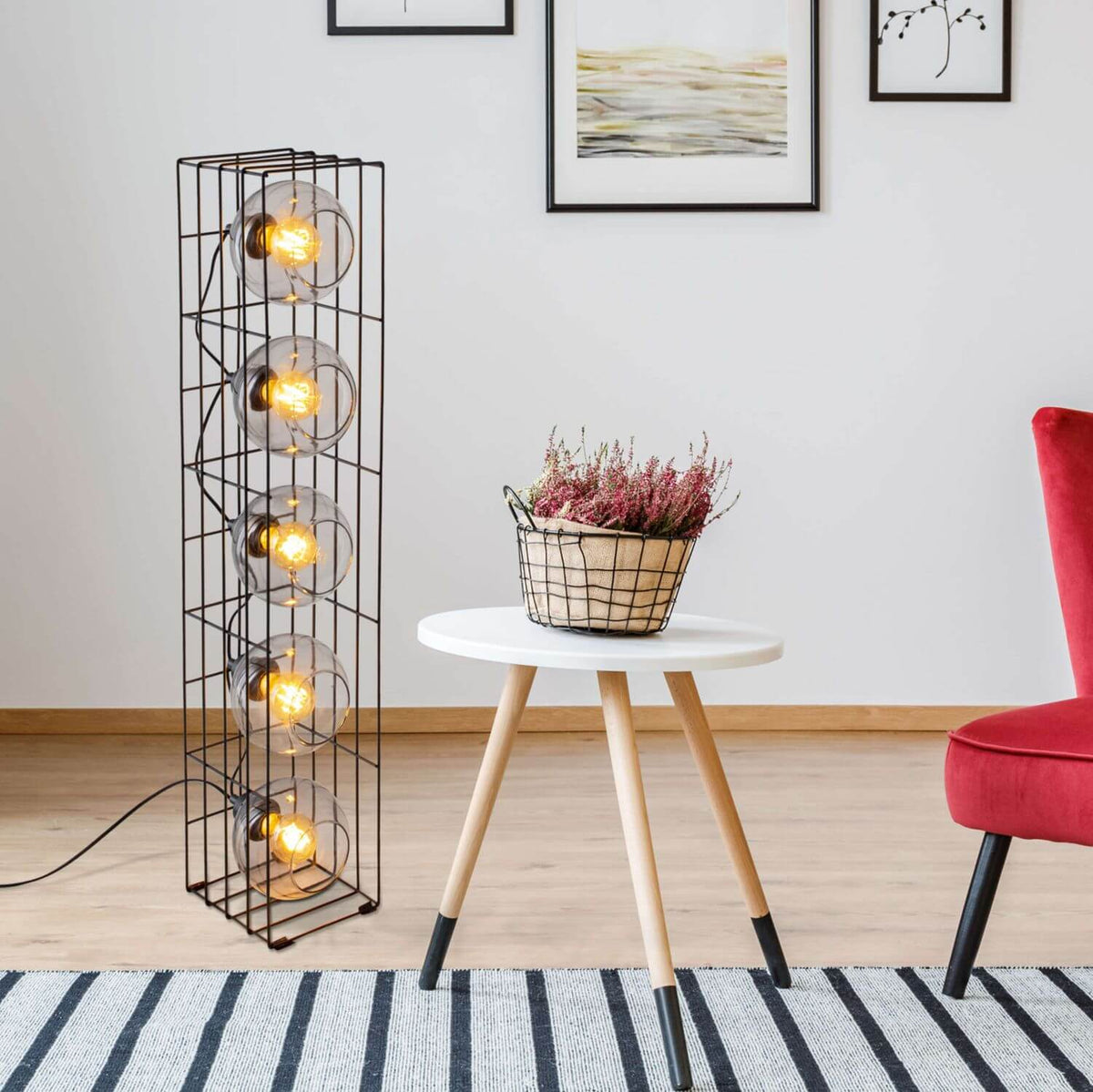 Lampadaire allumé dans un salon avec table blanche, plante, chaise rouge et tapis rayé.
