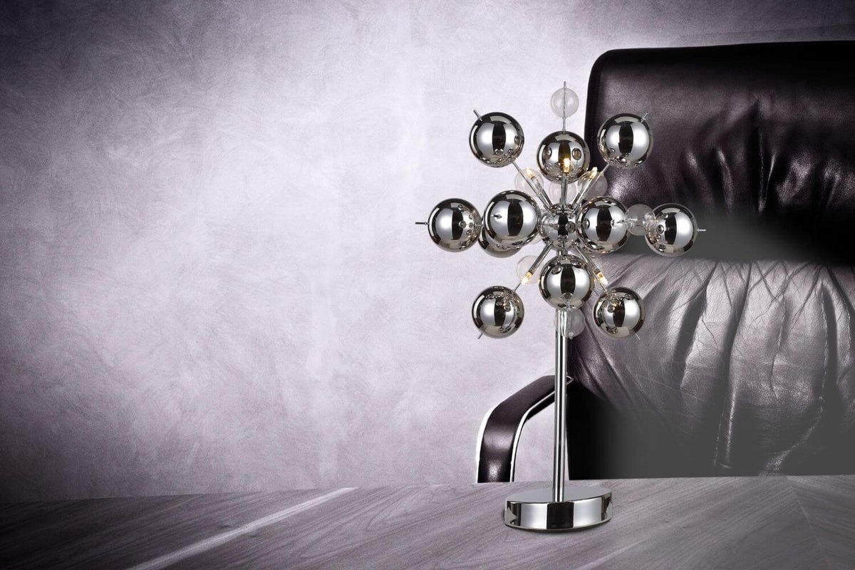 Mise en ambiance avec lampe chromée posée sur table en bois devant fauteuil noir.