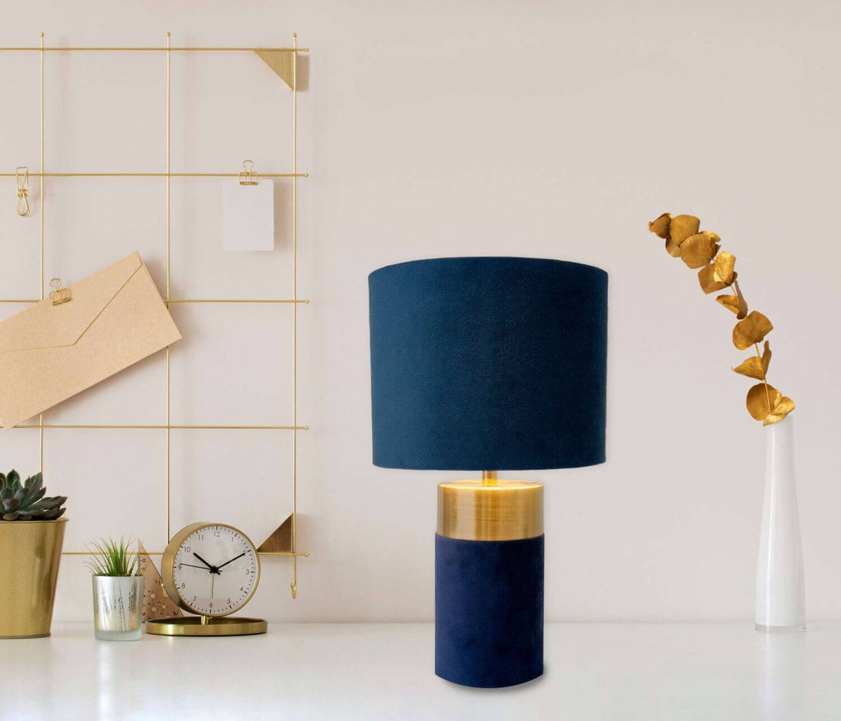 Lampe bleue posée sur un bureau blanc, entourée d'accessoires dorés et d'une plante verte.