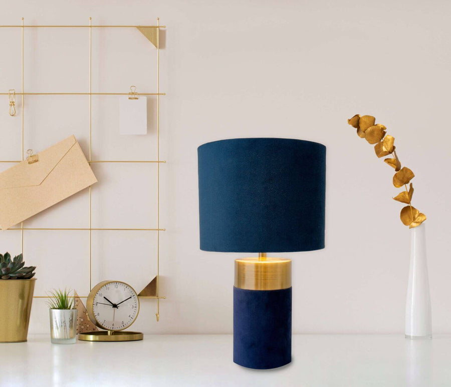 Lampe bleue posée sur un bureau blanc, entourée d'accessoires dorés et d'une plante verte.