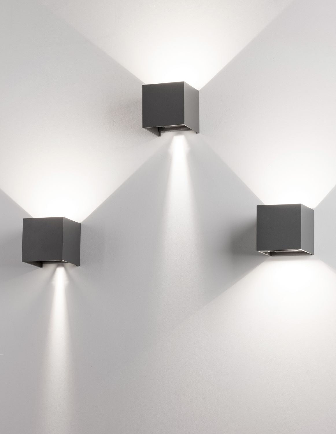 Mise en ambiance de trois appliques cubes éclairant un mur blanc, lumière rasante.