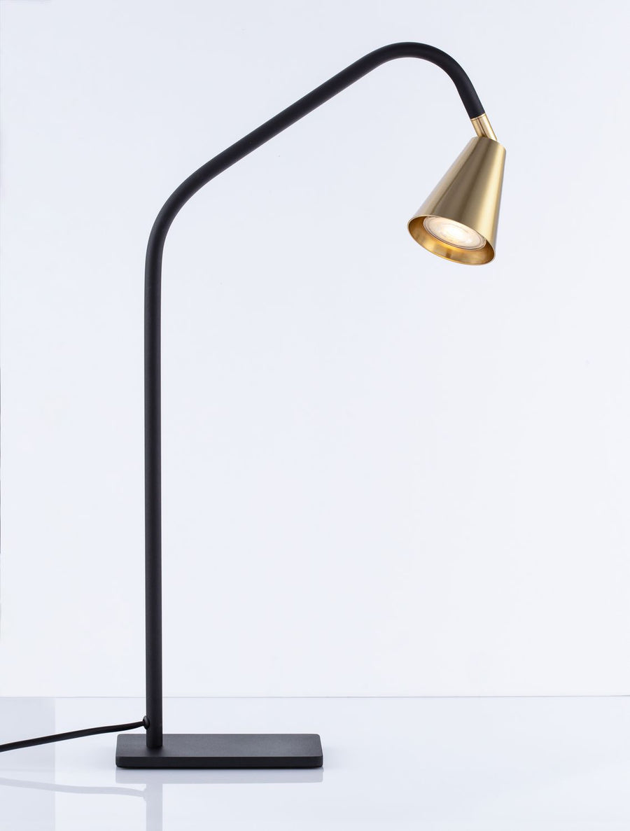 Lampe à poser complète allumée sur fond blanc, abat-jour conique laiton et tige noire.