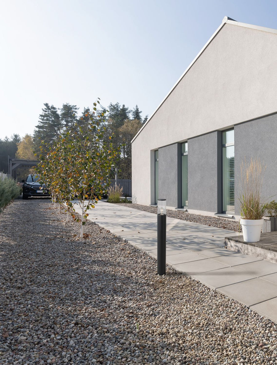 Borne noire installée dans un jardin de graviers devant une maison contemporaine, le long d'une allée.