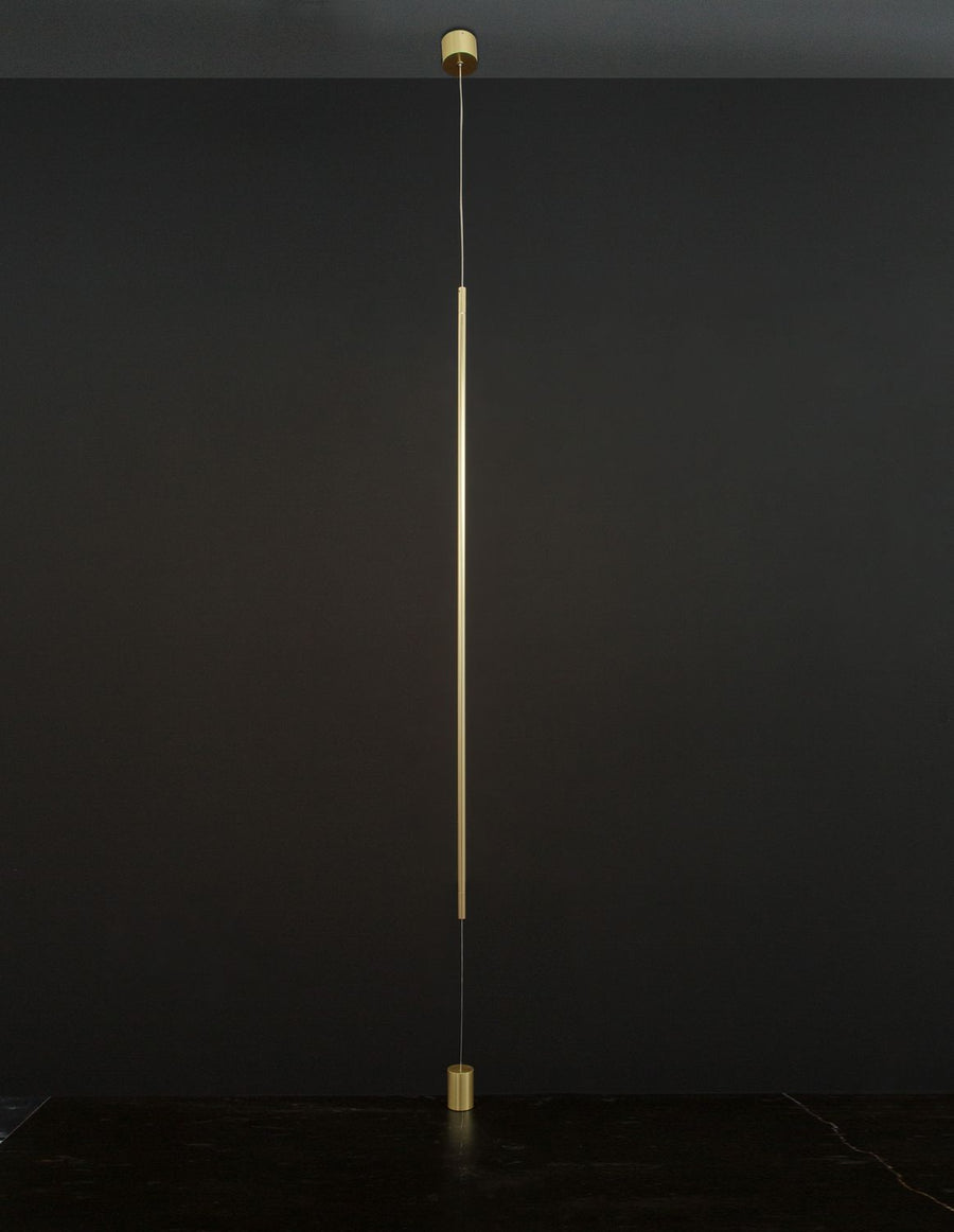 Suspension laiton installée devant un mur noir, éclairage vertical discret.