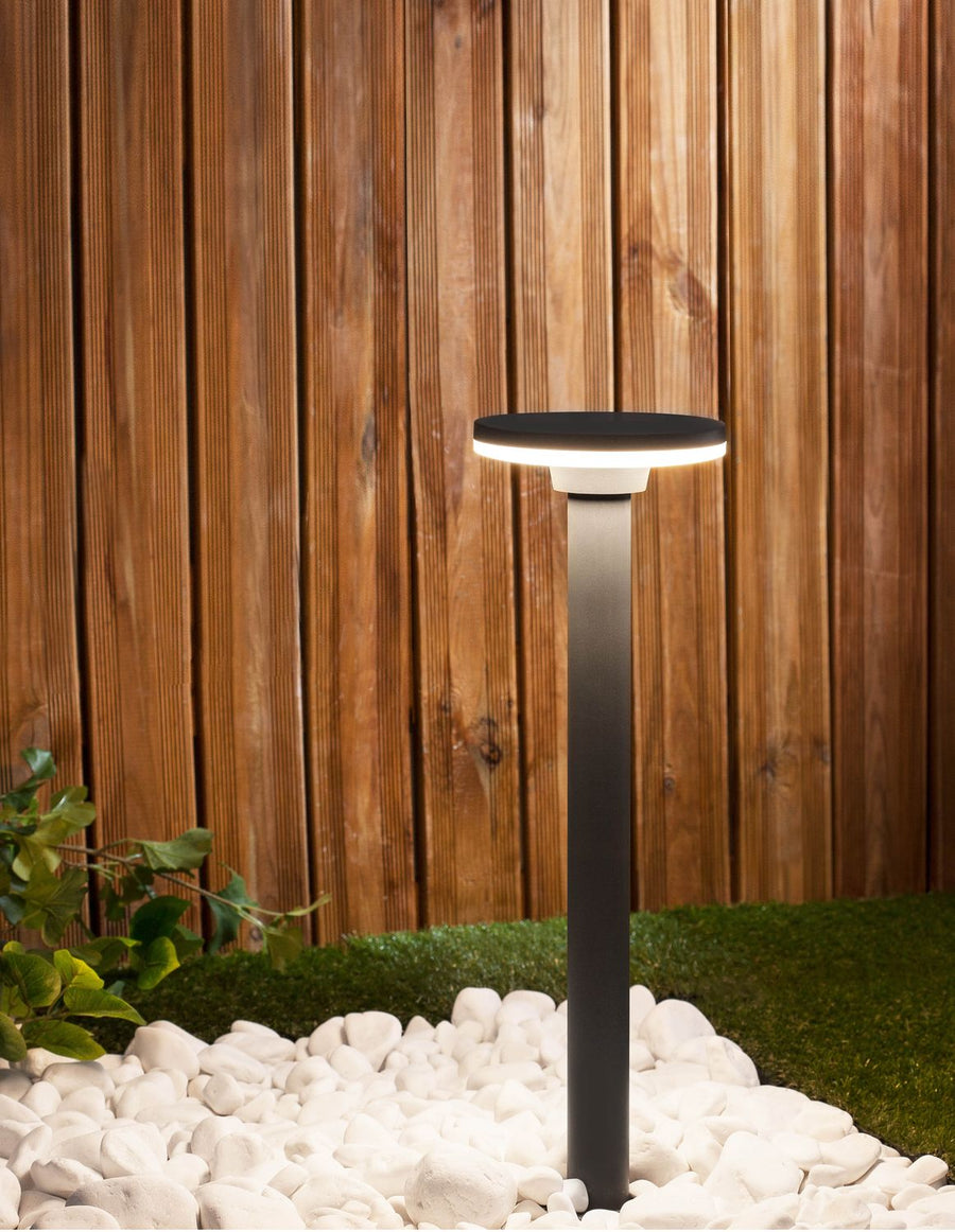 Borne lumineuse grise allumée dans un jardin, gazon, galets blancs et clôture en bois