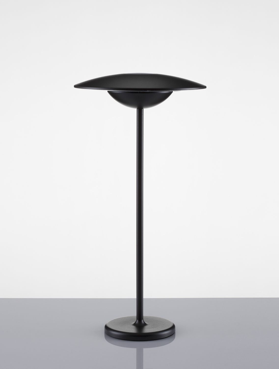 Luminaire noir de face posé sur sol gris réfléchissant, base ronde visible