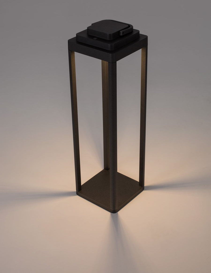 Lampadaire extérieur anthracite allumé sur fond beige, ombre portée au sol.