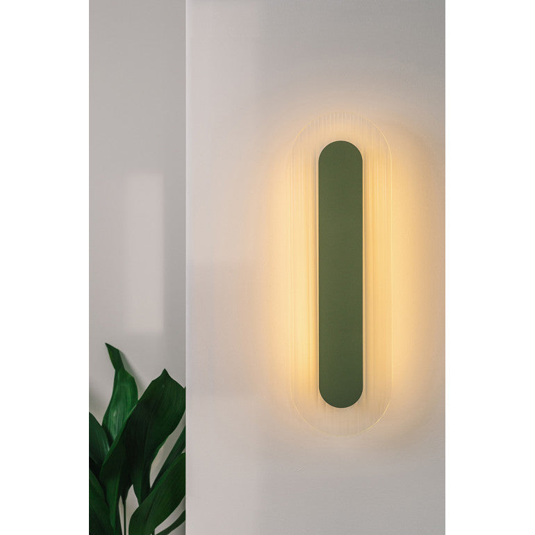 Mise en ambiance : applique murale allumée sur un mur clair, halo lumineux chaud et plante verte.