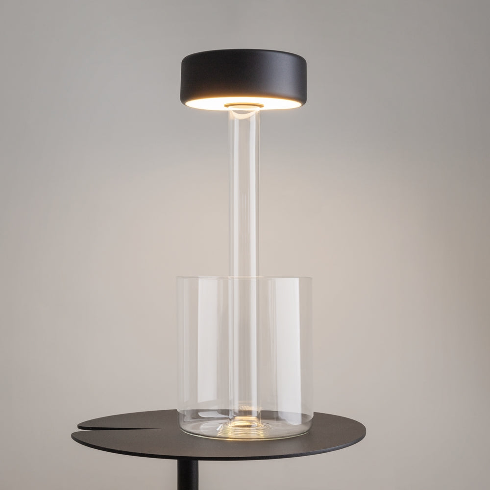 Lampe allumée posée sur table ronde noire, halo chaud, fond gris beige.