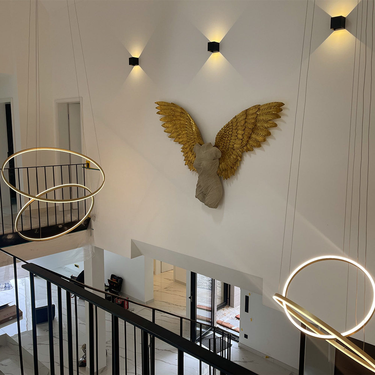 Deux luminaires annulaires dorés suspendus devant mur blanc et sculpture ailée dorée