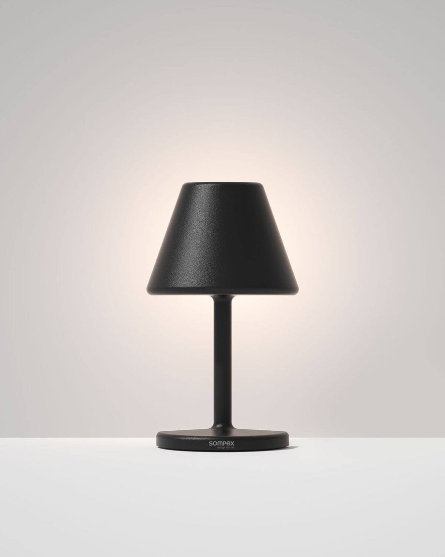Lampe noire allumée sur fond clair, texture granuleuse et base ronde.