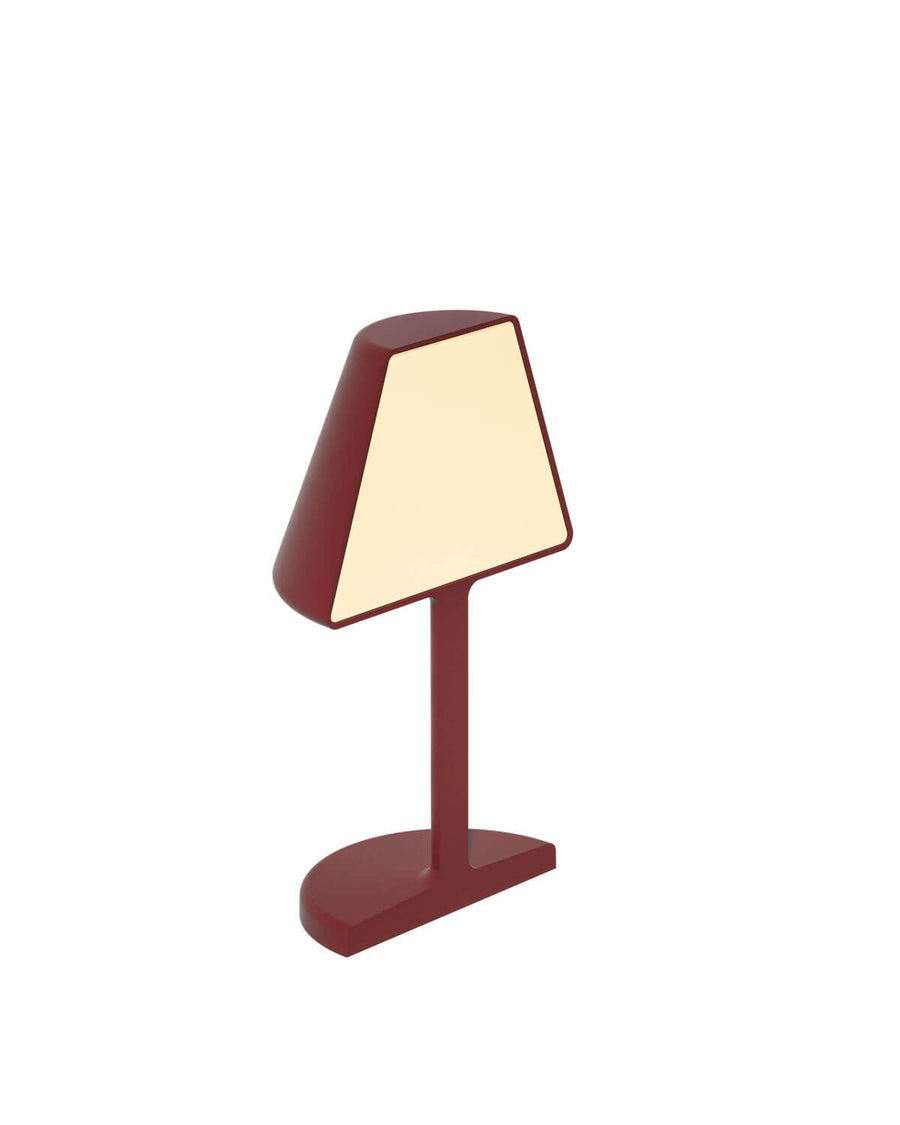 Vue de trois quarts du luminaire rouge, mettant en valeur l'abat-jour conique et le socle ovale.