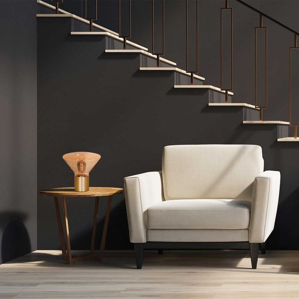 Lampe posée sur une table d'appoint près d'un fauteuil, sous un escalier au mur sombre.