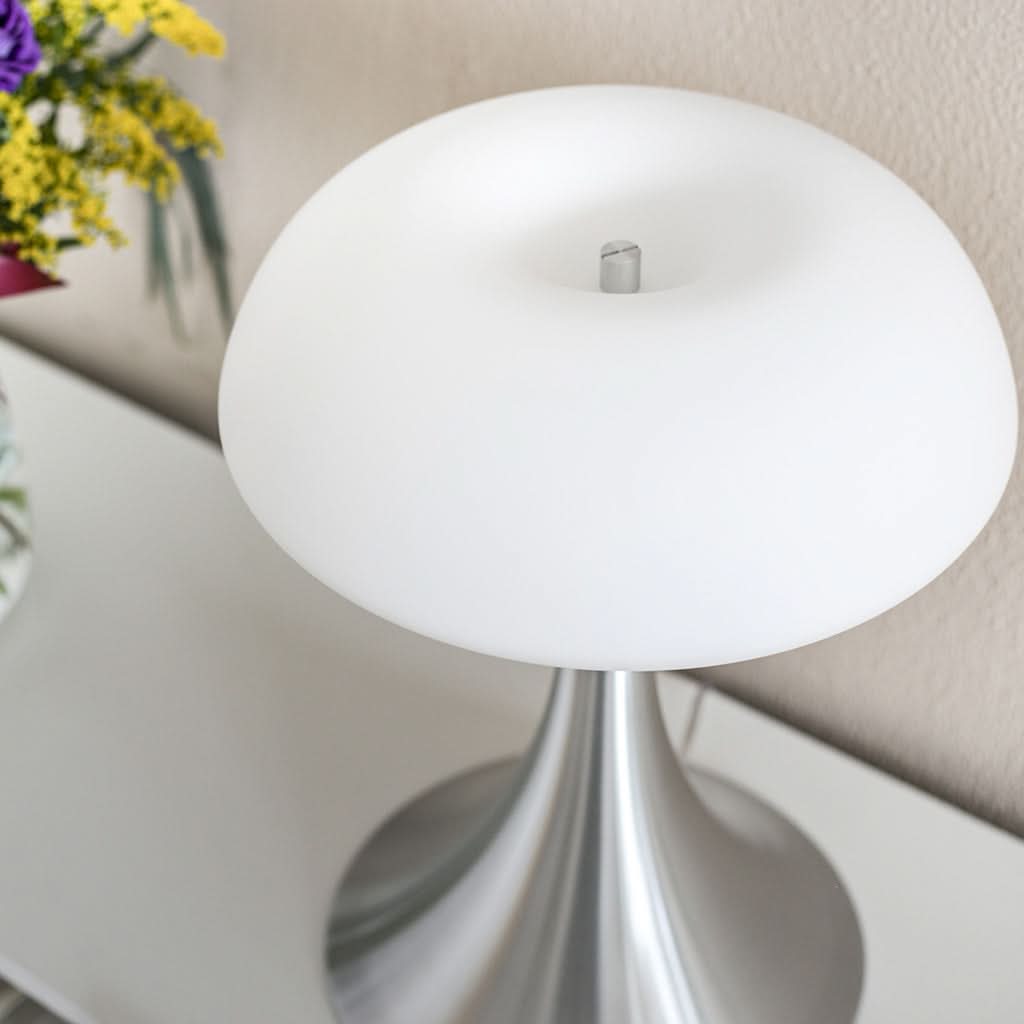 Lampe blanche et argent posée sur un meuble, fleurs floues en arrière-plan