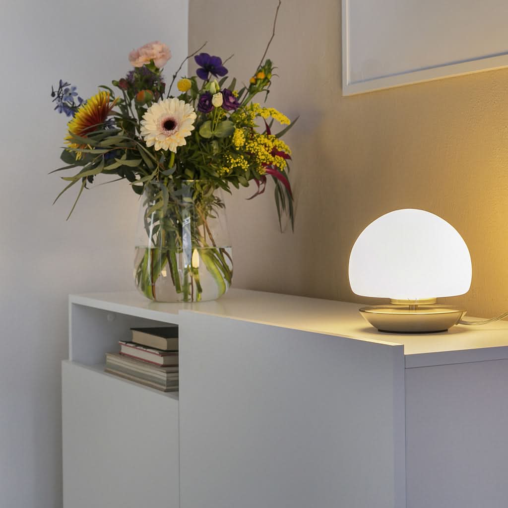 Mise en ambiance: lampe allumée sur meuble blanc, près d'un vase de fleurs et de livres.