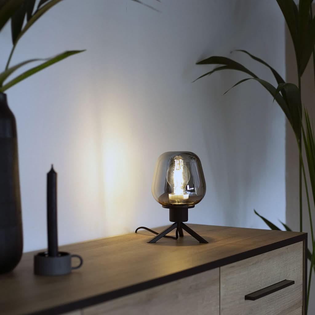 Ambiance tamisée, lampe de table allumée sur meuble en bois, lumière chaude projetée au mur.