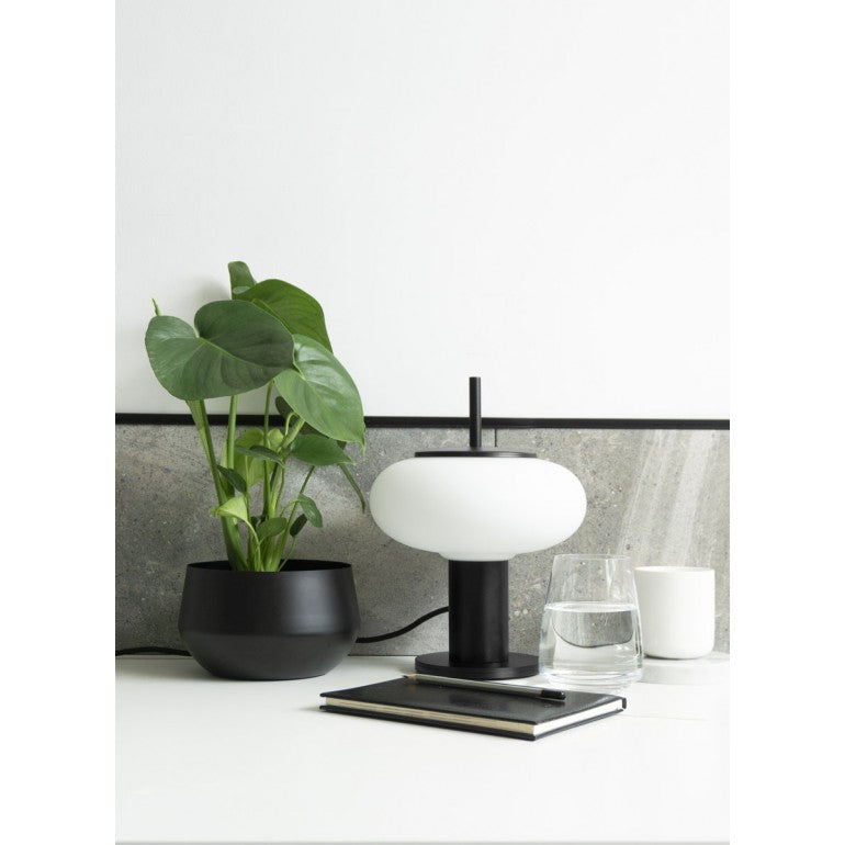 Lampe noire posée sur table avec plante, carnet, verre d'eau et mur gris.