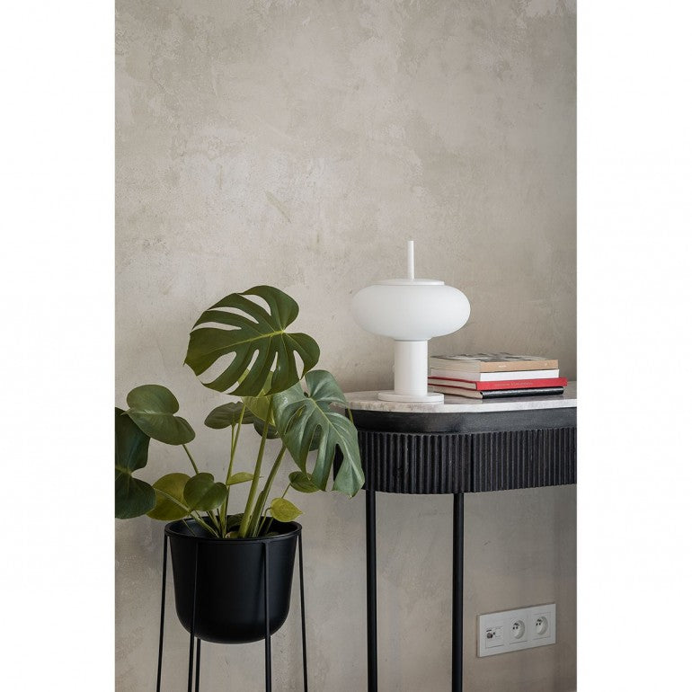 Lampe blanche en situation sur table d'appoint, plante verte, livres et prise murale.