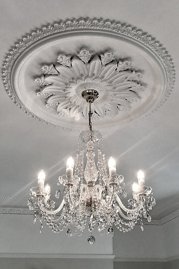 Vue en contre-plongée du lustre sous rosace décorative, détails cristal et lumière blanche.