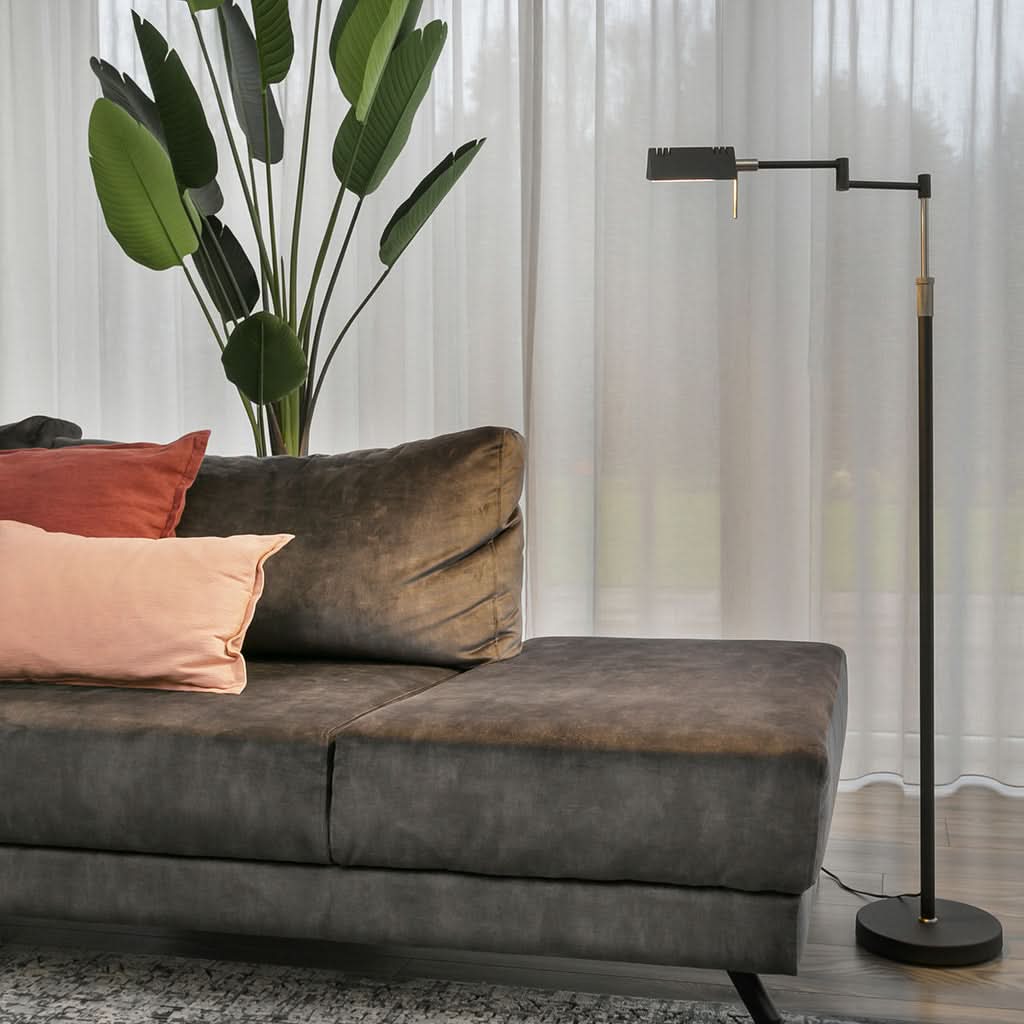 Lampe sur pied noire allumée devant un canapé en velours et rideaux clairs.