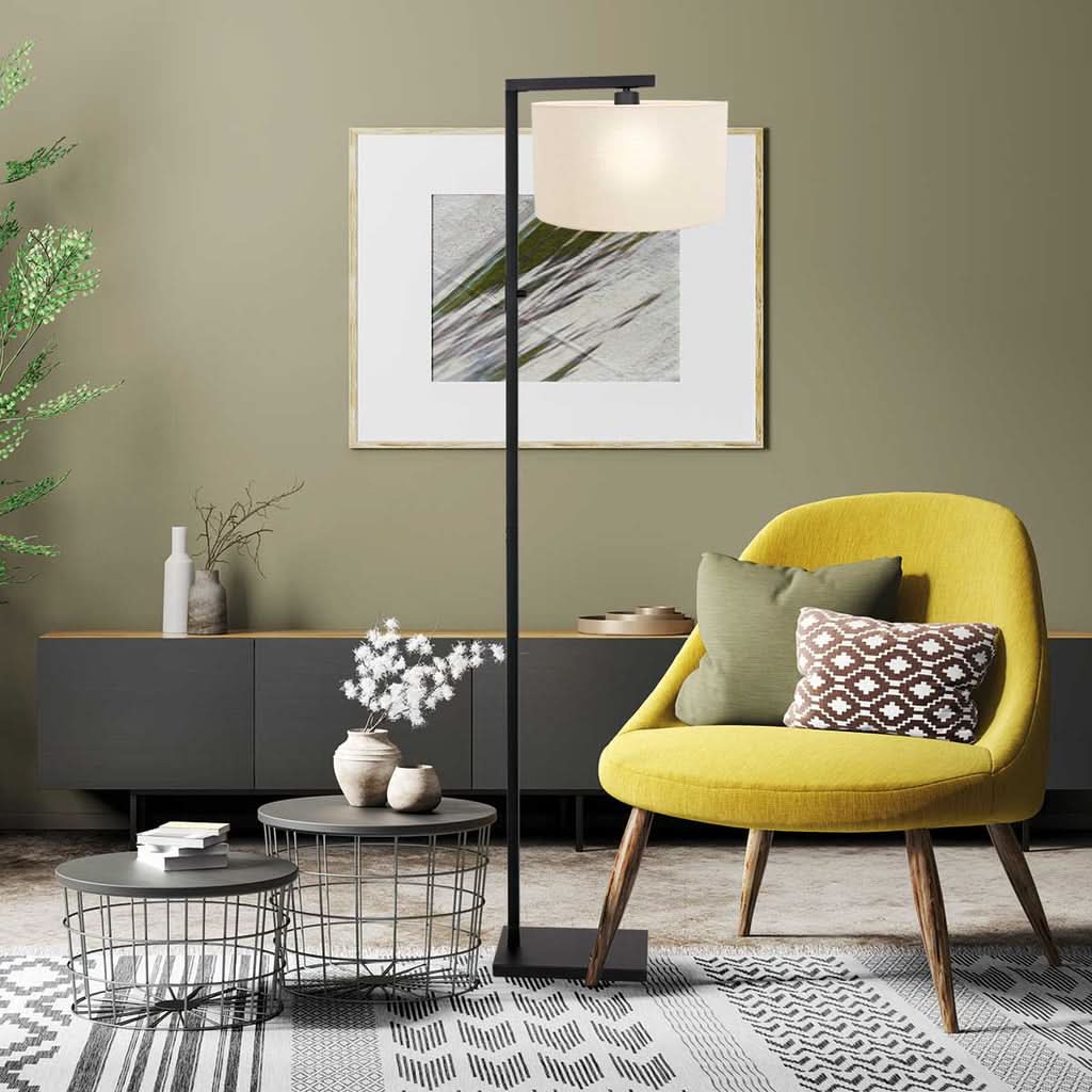 Mise en ambiance salon avec lampadaire noir allumé, abat-jour cylindrique blanc et fauteuil jaune.