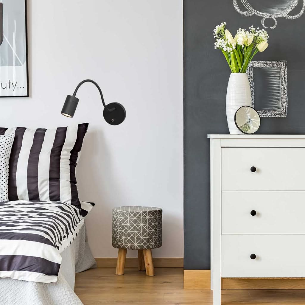 Applique murale noire allumée au-dessus d'un lit, dans une chambre avec coussins rayés et commode blanche.