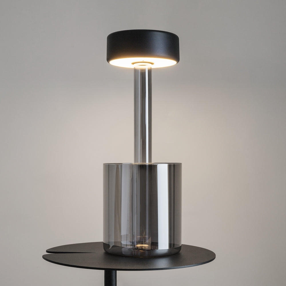 Lampe de chevet grise allumée posée sur table, verre fumé et halo lumineux chaud.