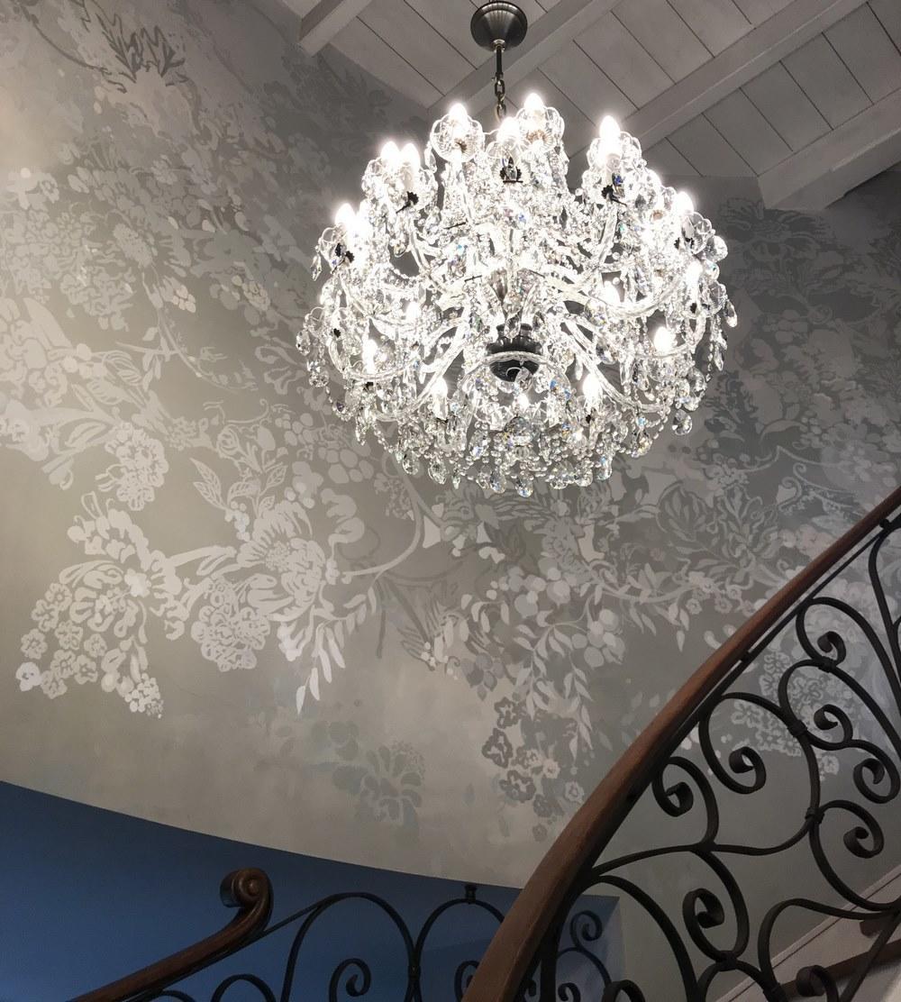Lustre allumé en hauteur dans cage d'escalier, papier peint floral et rampe en fer forgé.