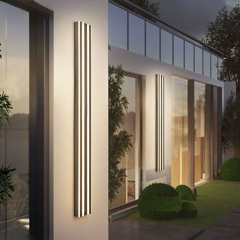 Façade moderne avec plusieurs appliques linéaires dorées allumées, reflets dans les baies vitrées et jardin.