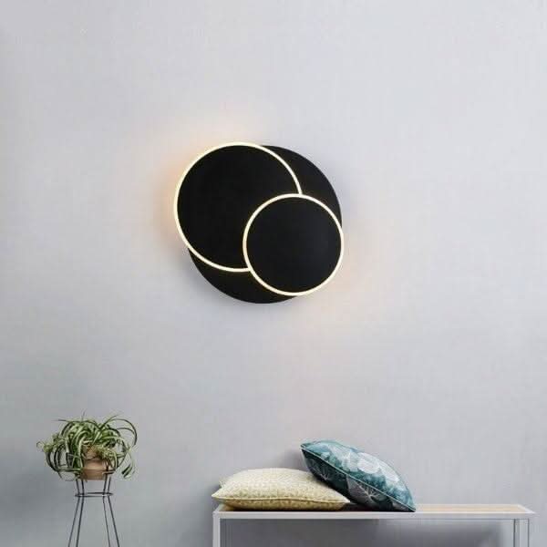 Mise en ambiance d'une applique murale noire allumée sur un mur gris, avec meuble et coussins